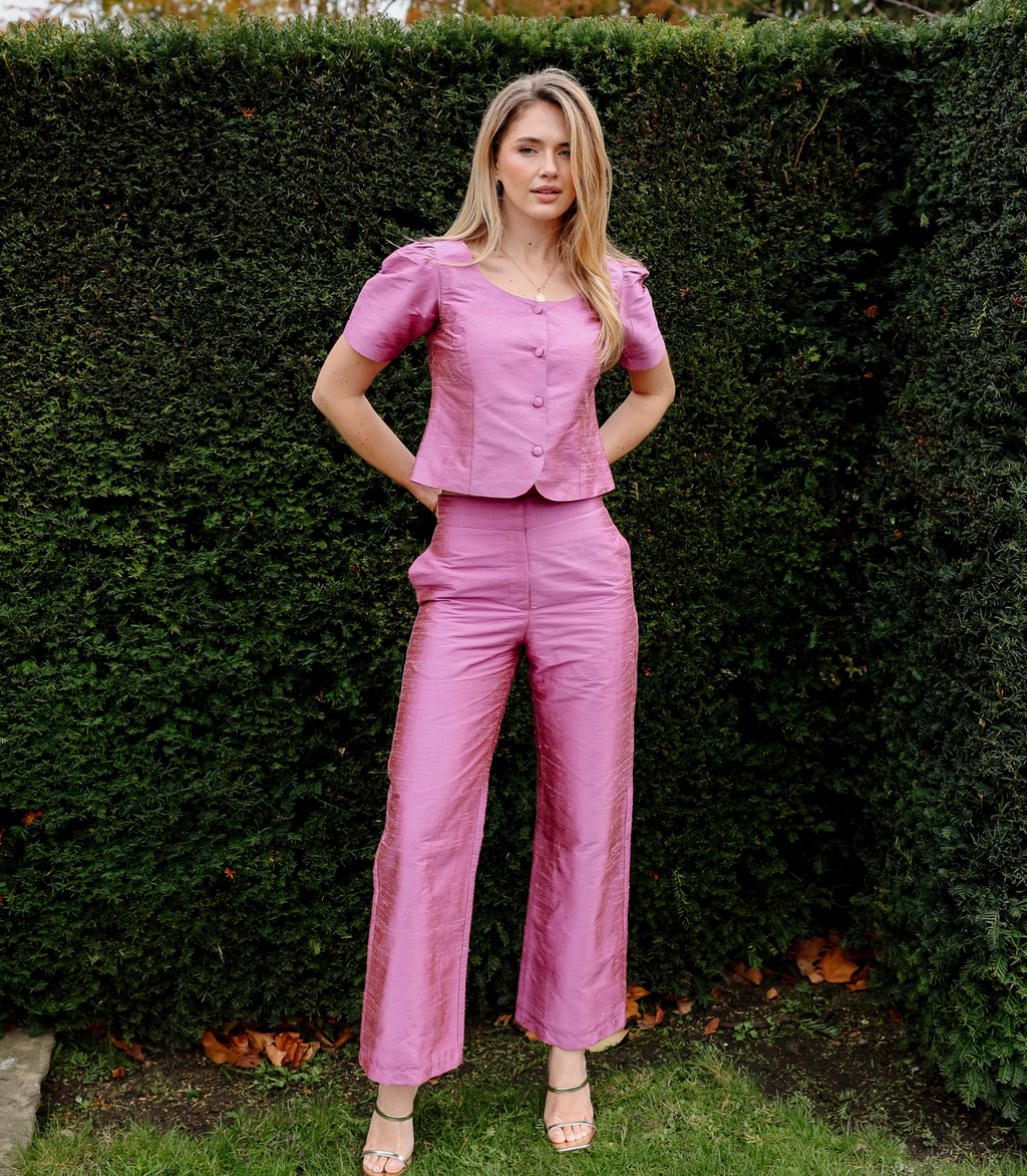 A woman wearing the Jane Top in Lychee, a pink short-sleeve button-down top with a square neckline, paired with matching wide-leg trousers.