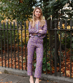 Full-length view of a woman wearing the Harper tailored trousers in a purple lilac and yellow goldenberry floral jacquard pattern, standing by a garden fence.