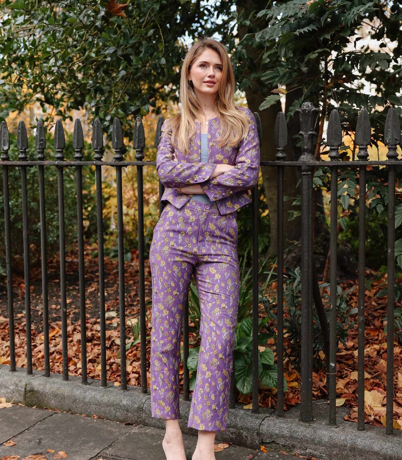 Full-length view of a woman wearing the Harper tailored trousers in a purple lilac and yellow goldenberry floral jacquard pattern, standing by a garden fence.
