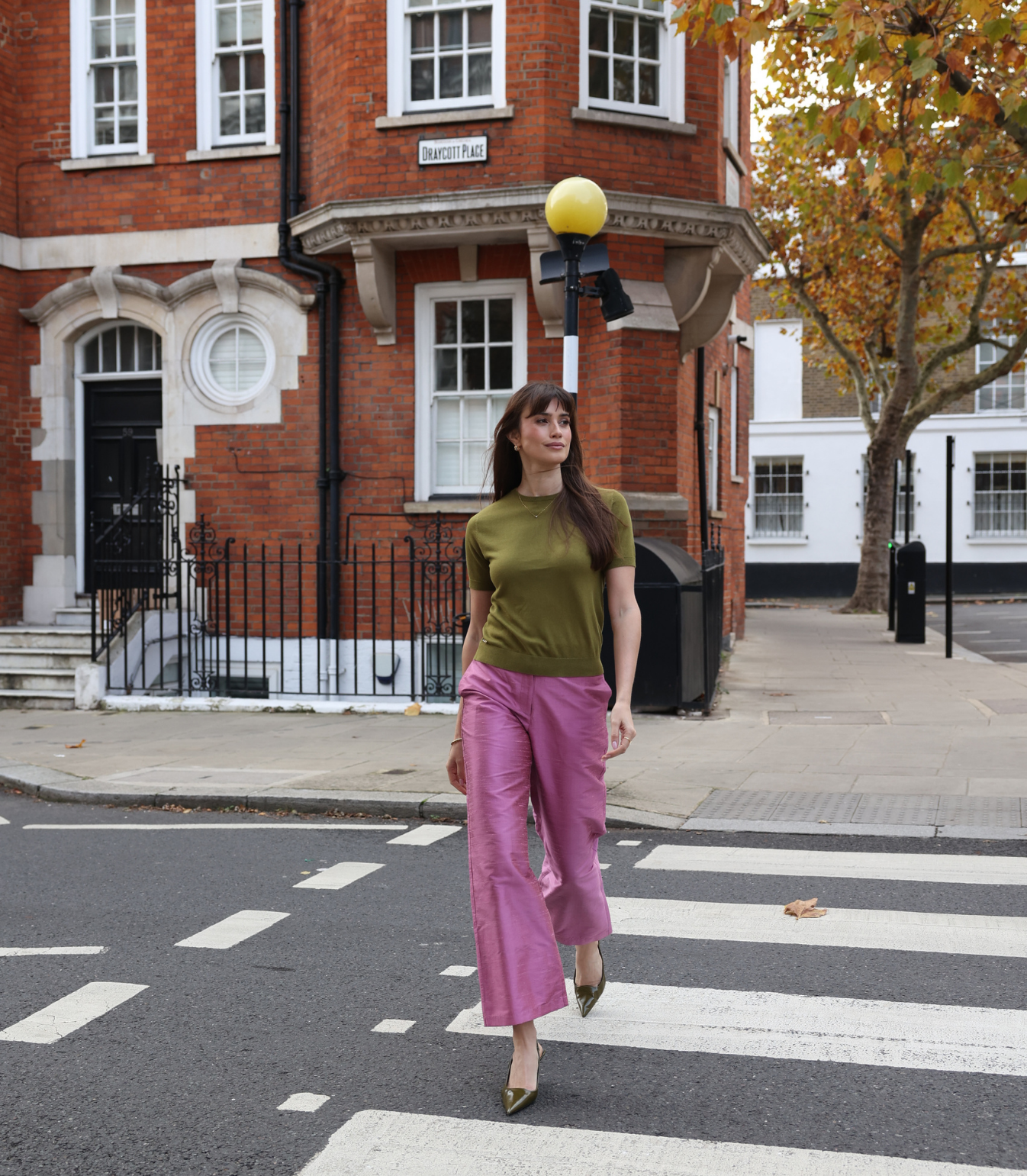 A woman walking in the city wearing an olive green short-sleeve knit top and magenta wide-leg trousers, showcasing a versatile street-style look.