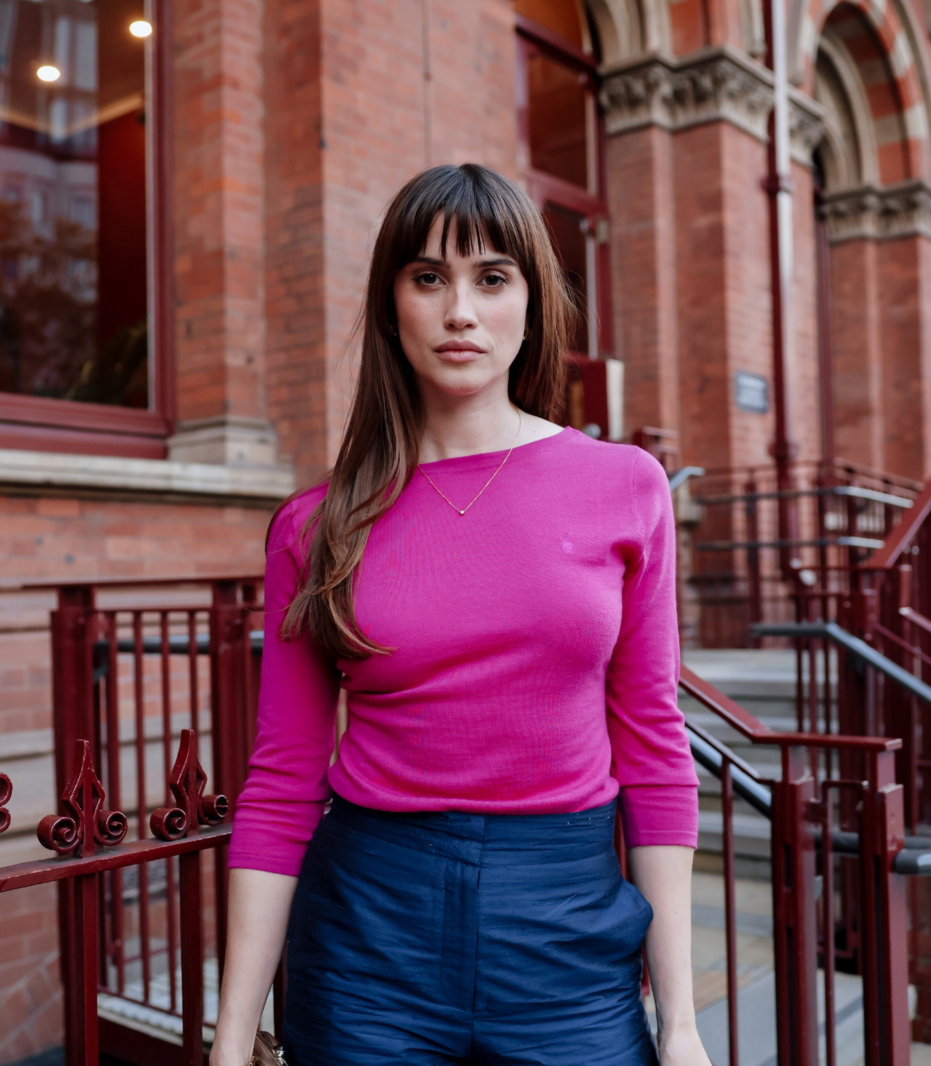 Woman wearing a bright pink sweater and blue pants standing in front of a brick building.